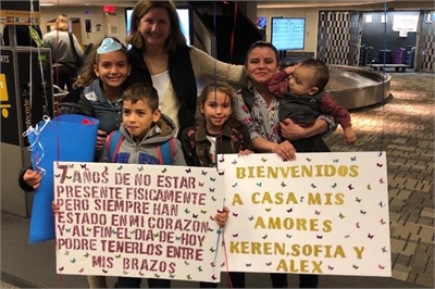 [ai] A group of five people, including a woman holding a child and four children, stands at an airport's baggage claim area. They are holding signs welcoming family members, with one sign in Spanish expressing love and reunion after several years.